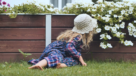 Young child resting on grassy ground, wearing handmade crocheted hat, gazing at white petunia blooms in wooden raised garden bed, relaxing outdoorsの写真素材