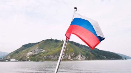 Russian flag waving from a boat sailing on secluded lake baikal, with a green hill and cloudy sky in...