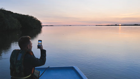 Fisherman wearing life jacket taking pictures of beautiful sunset over calm lake from boat, enjoying...