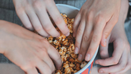 Hands grabbing caramel popcorn during sunny park gathering, friends sharing tasty treat outdoorsの写真素材