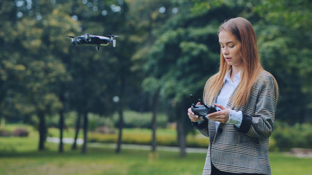Businesswoman piloting a drone in a park, holding a remote control and navigating the aircraft...