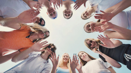 Group of young students forming a joyful circle, raising their hands against a bright, clear sky, celebrating friendship and unityの写真素材