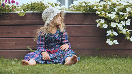 Barefoot girl with hat sitting on grass near wooden fence and white petunias, enjoying family day in gardenの写真素材