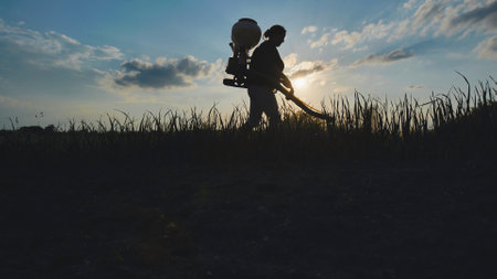 Farmer walking on cultivated field spraying pesticides with manual knapsack sprayer at sunsetの写真素材