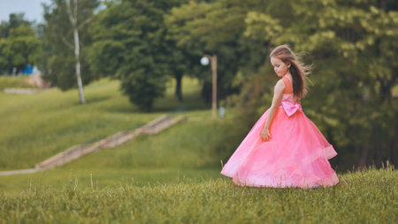 Young girl in pink dress walking cheerfully across verdant park, experiencing summer sunlight and natural surroundingsの写真素材