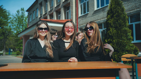Three young women wearing trendy sunglasses, sitting together at a table outside their school,...