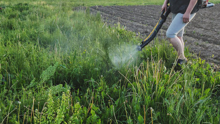 Farmer spraying pesticides with backpack sprayer near plowed field rows, targeting weeds