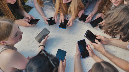Group of teenagers lying on grass using smartphones together around white table.