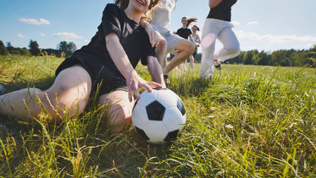 Group of young adults is running after a soccer ball on a grass field on a sunny summer day.