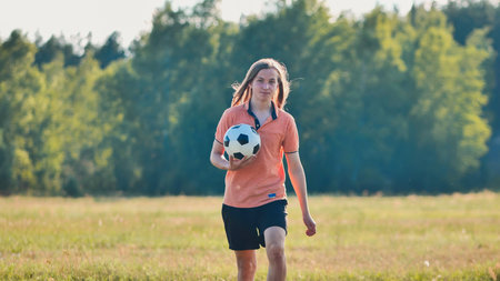 Young woman is walking through a field on a sunny summer day, holding a soccer ball after a casual game with friends.の写真素材