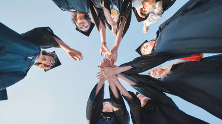 Young graduates in caps and gowns come together in a circle to celebrate their achievement by joining handsの写真素材