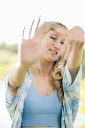 Blonde woman framing face with hands, expressing playful mood while standing outdoors in summer casual attireの写真素材