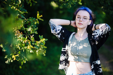 Gen z student with dyed blue hair wearing glasses and casual clothes relaxing in a park under oak...