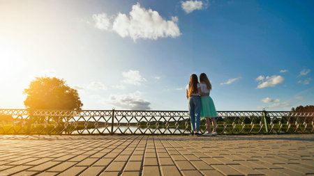 Two school girlfriends embracing on a bridge, sharing laughter and joy while admiring a stunning...