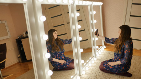 The girl sits on the floor and makes selfies at the makeup mirrors at home.の写真素材