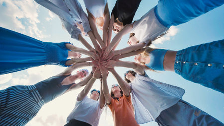 School friends standing in a circle, linking hands against a blue sky, representing unity, teamwork, and collaborationの写真素材