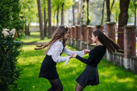 Two cheerful schoolgirls, wearing black skirts, happily twirling and holding hands on a lively green schoolyard grassの写真素材