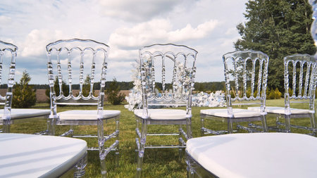 Elegant transparent chairs with white cushions arranged on lawn for outdoor wedding ceremony under cloudy skyの写真素材