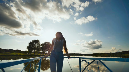 Girl with long hair, wearing denim overalls, standing on a pier, fishing rod in hand, enjoying a sunny day by the lakeの写真素材