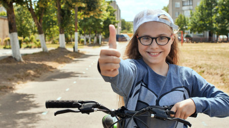 Smiling schoolgirl with glasses and cap giving thumbs up while pedaling bicycle under bright sunlightの写真素材