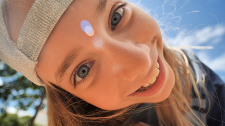 Cheerful schoolgirl sporting white baseball cap, beaming with vibrant smile and sparkling blue eyes under sunny skyの写真素材
