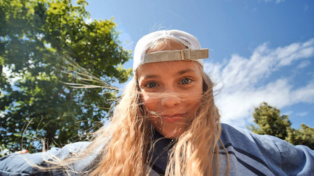 Cheerful schoolgirl in cap, wind tousling hair, basking in bright sunlight while playing outdoors in...