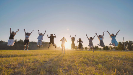 High school students jubilantly leaping with arms raised in a field at sunset, relishing their summer vacationの写真素材