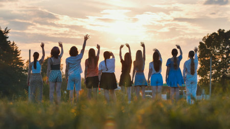 Students happily waving goodbye in a field against a beautiful sunset, celebrating the end of school and the beginning of summerの写真素材
