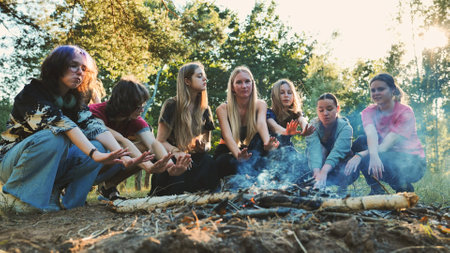 Young students huddling near crackling campfire, warming hands and sharing camaraderie during woodland camping adventureの写真素材