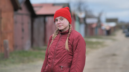 Confident teenage girl wearing red beanie, glasses, jacket, standing near colorful beach huts with relaxed poseの写真素材