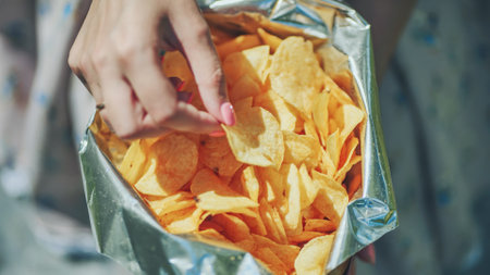 Woman hand reaching into metallic foil bag, grasping crunchy potato chip with anticipation of salty, crisp flavorの写真素材