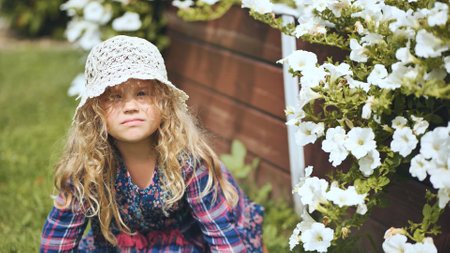 Cheerful child wearing handmade crochet hat smiling among blooming white petunias during family garden gatheringの写真素材