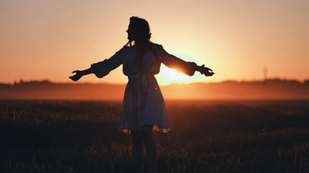 Woman silhouetted, arms spread wide, embracing golden wheat field sunset with joyful sense of liberationの写真素材