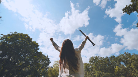 Young musician raises her clarinet and arms in triumph, celebrating a successful performance in a lush park under a bright blue skyの写真素材