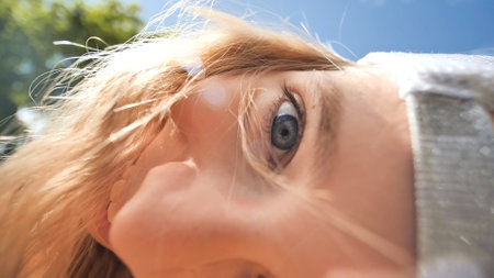Blonde schoolgirl tilting head, windswept hair framing blue eye against azure sky backdropの写真素材