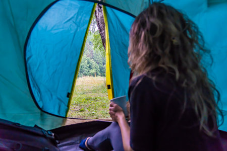Young woman sitting in her tent, holding a mug and looking at the landscape, enjoying the quiet of natureの写真素材