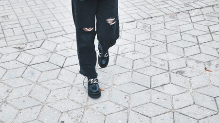 Trendy female legs striding across geometric concrete pavement, wearing distressed black jeans and chunky chain adorned bootsの写真素材