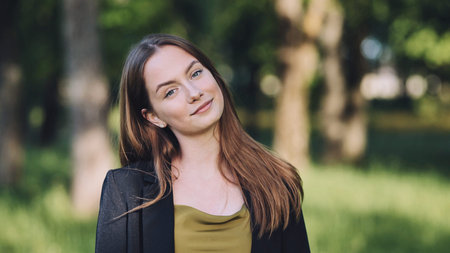 Portrait of a confident businesswoman smiling slightly in a park, enjoying the fresh air and...