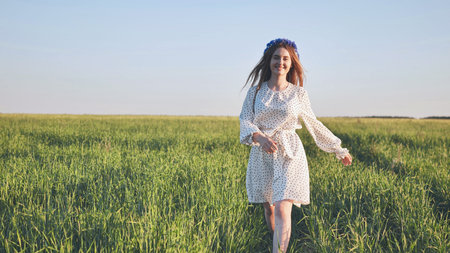 Joyful woman wearing floral headpiece running across verdant meadow under sunny skyの写真素材