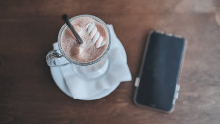 Hot chocolate with fluffy marshmallows resting near smartphone on rustic wooden surface, conveying warm, relaxing momentの写真素材