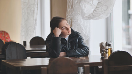 Young woman sitting alone at a table in a restaurant, gazing out the window with a bored expression while waiting for someoneの写真素材