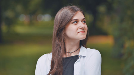 Portrait of a cheerful young woman with long brown hair, wearing a white shirt, smiling and looking up in a parkの写真素材