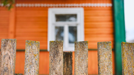 Weathered wooden fence in front of an orange wooden house with a white window, creating a rural and nostalgic atmosphereの写真素材