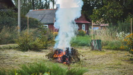 Smoldering tree branches creating dense smoke plume near wooden house, rural garden setting with rustic wooden fenceの写真素材