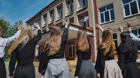 Female students happily waving goodbye in front of their school building, commemorating their accomplishments on a sunny dayの写真素材