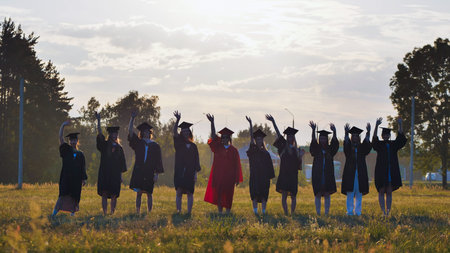 Happy graduates in graduation gowns waving at sunset in a field after ceremony