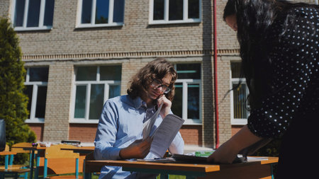Two students are studying together at a desk in a schoolyard, preparing for exams on a sunny day