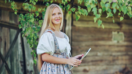 Young blonde woman wearing traditional slavic dress using digital tablet in rural landscape near old wooden houseの写真素材