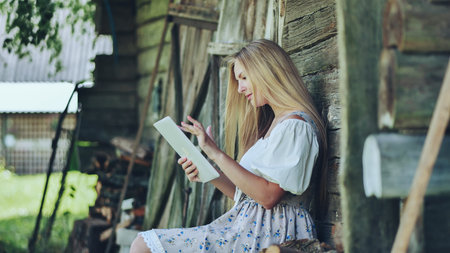 Blonde woman browsing digital tablet, sitting in pastoral landscape with rustic wooden fence and wildflower meadowの写真素材
