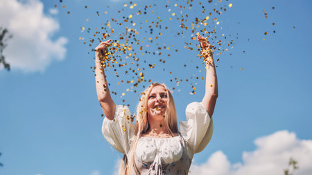 Happy caucasian woman throwing golden confetti in the air against blue sky, celebrating achievement...